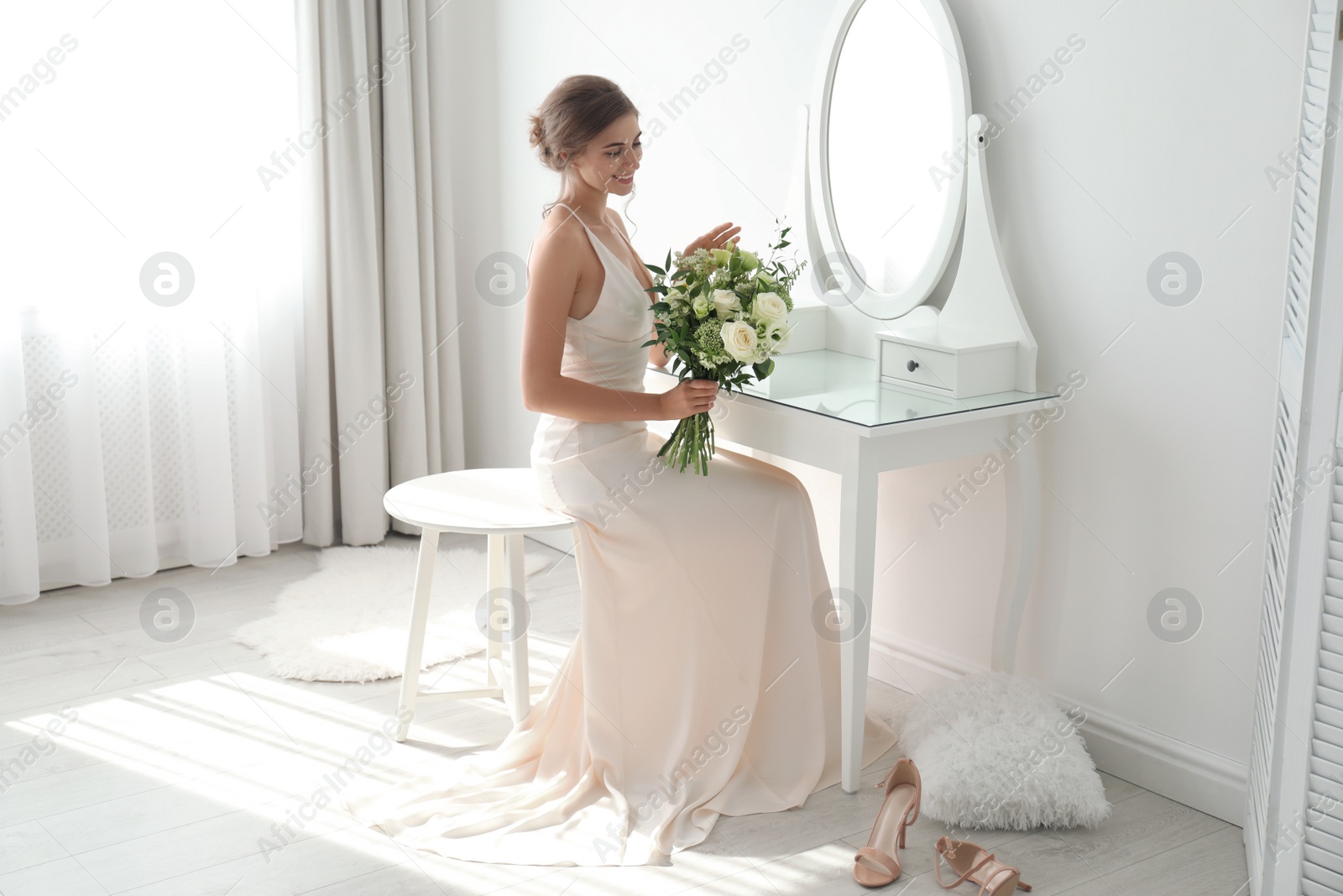 Photo of Young bride in wedding dress with beautiful bouquet near mirror indoors