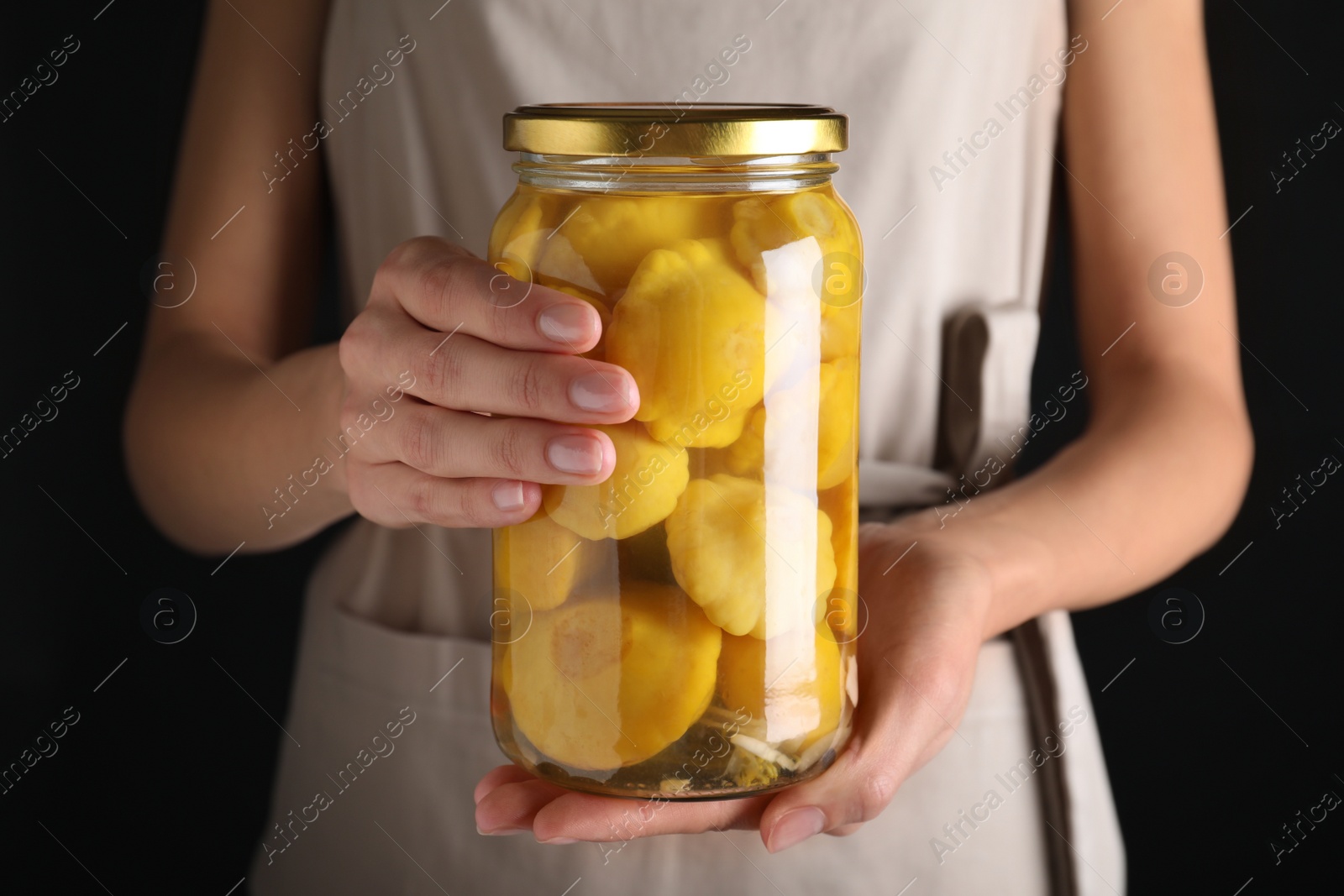 Photo of Woman holding jar with pickled pattypan squash against black background, closeup