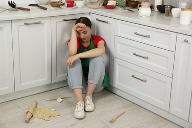 Photo of Tired woman sitting on dirty floor with utensils and food leftovers in messy kitchen