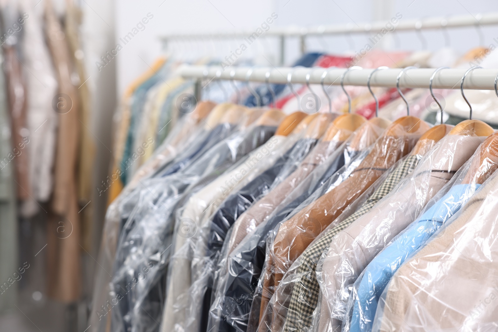 Photo of Dry-cleaning service. Many different clothes in plastic bags hanging on rack indoors, closeup