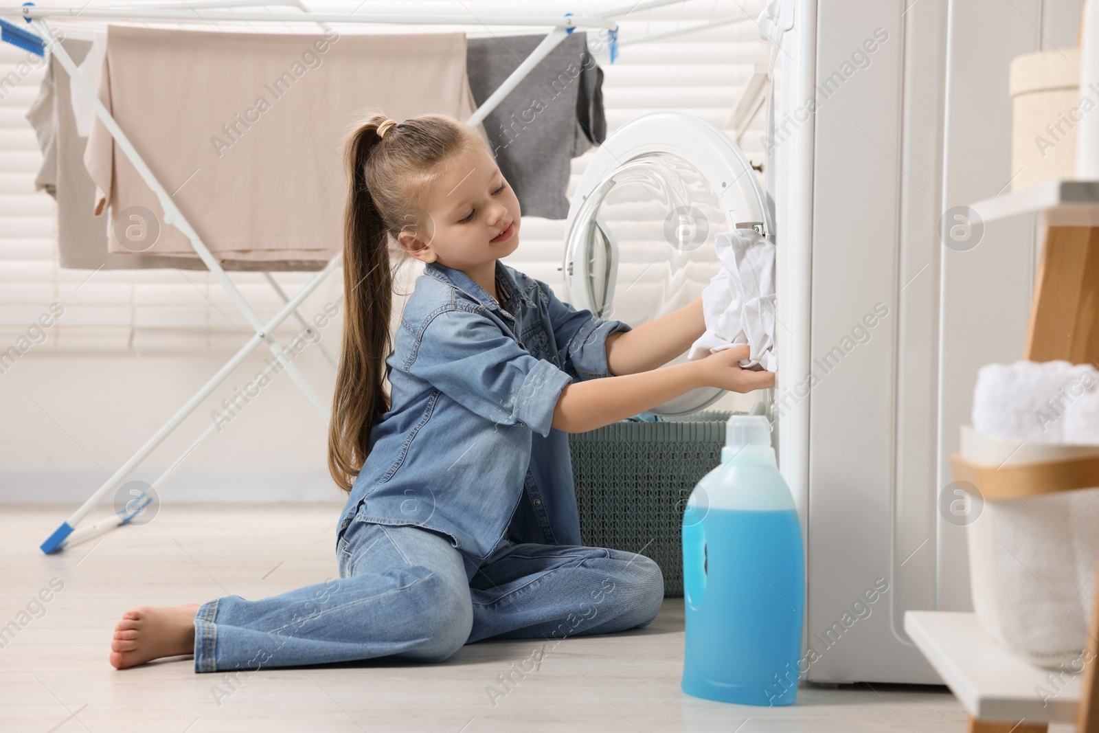 Photo of Little girl putting dirty clothes into washing machine in bathroom