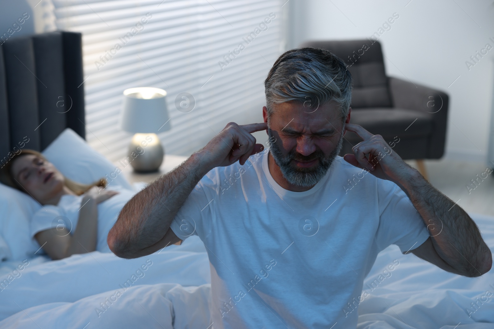 Photo of Irritated man covering his ears in bed at home, space for text. Problem with snoring wife