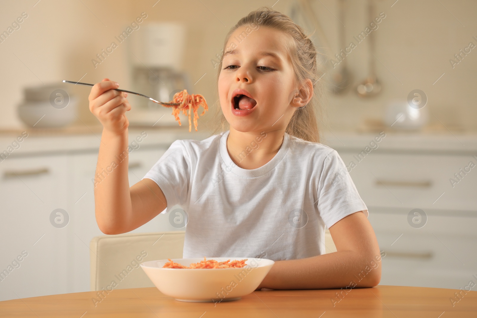 Photo of Cute little girl eating tasty pasta at table in kitchen