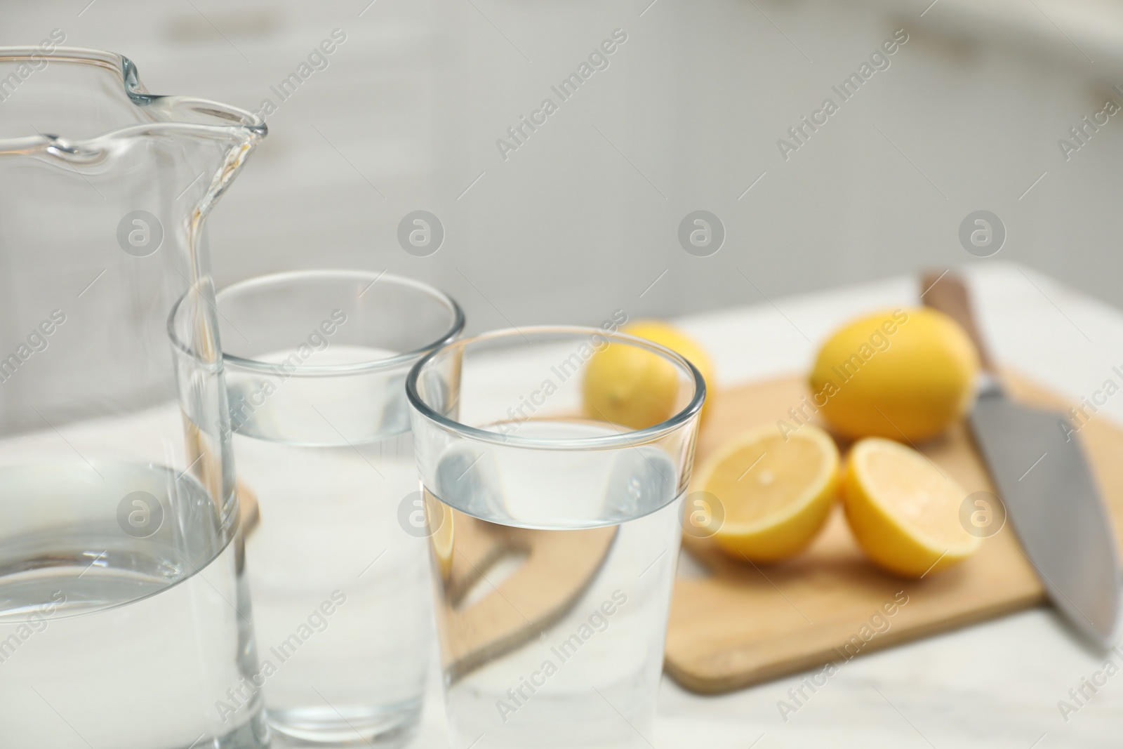 Photo of Jug, glasses with clear water and lemons on white table in kitchen, closeup