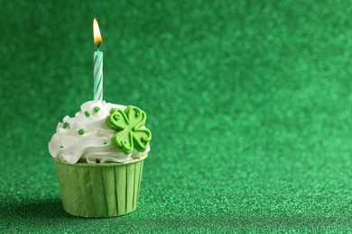 Photo of St. Patrick's day party. Tasty cupcake with clover leaf topper and burning candle on shiny green background. Space for text