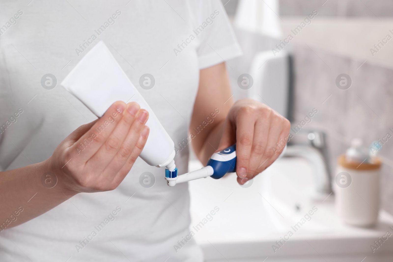 Photo of Woman squeezing toothpaste from tube onto electric toothbrush in bathroom, closeup