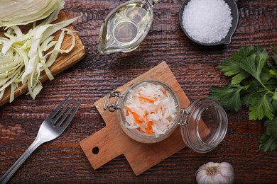 Glass jar of tasty sauerkraut and ingredients on wooden table, flat lay