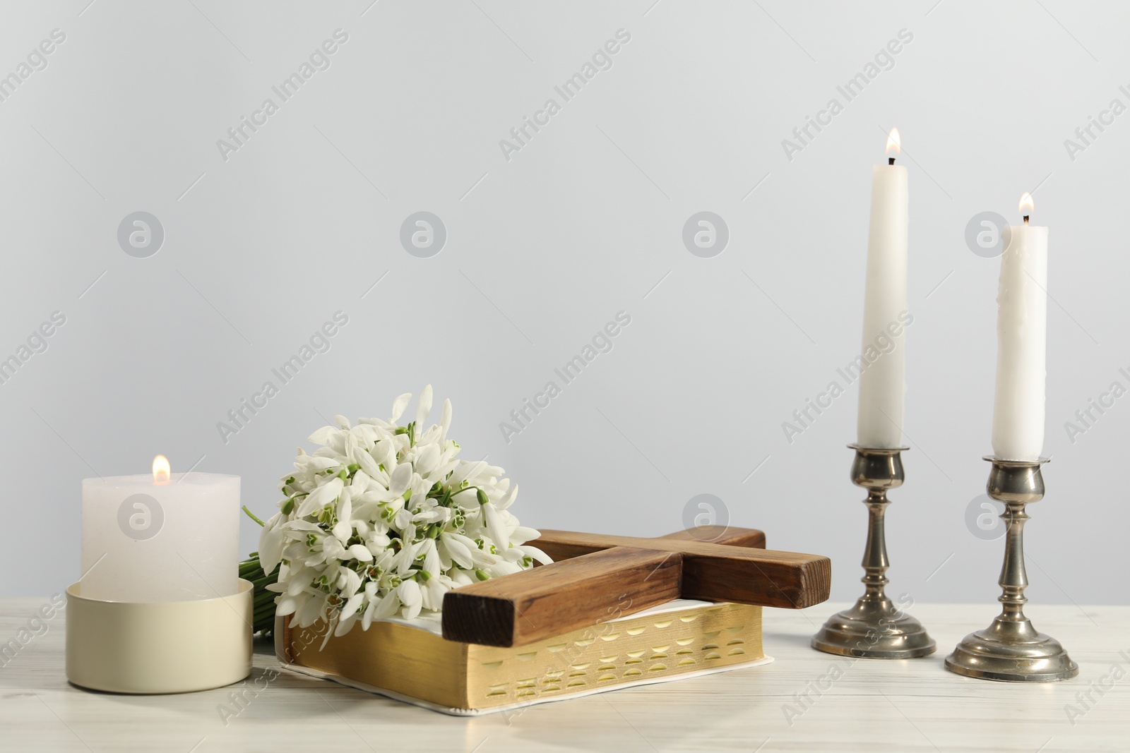 Photo of Burning church candles, wooden cross, Bible and flowers on white table