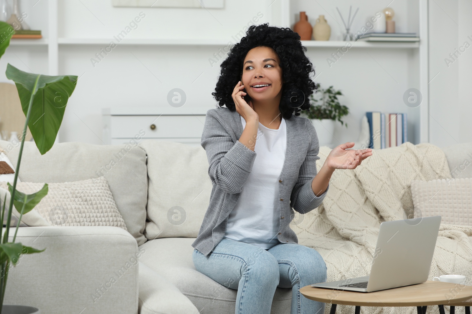 Photo of Happy young woman talking on phone while using laptop at coffee table indoors. Space for text