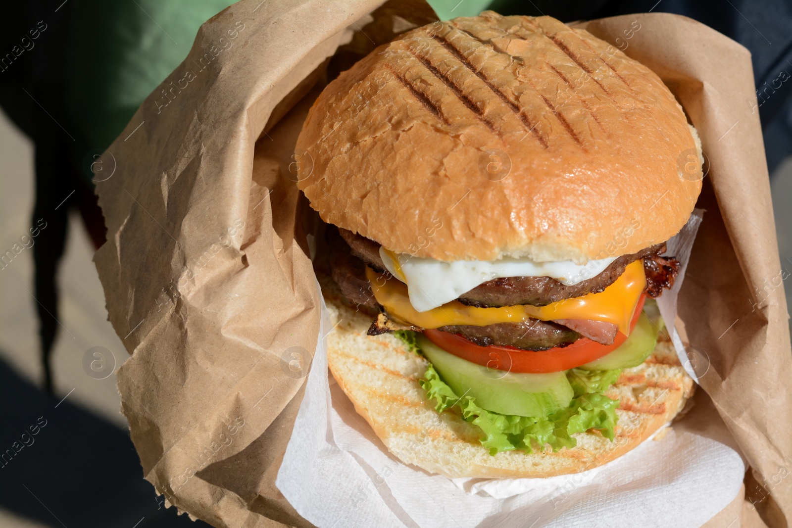 Photo of Woman holding delicious burger in paper wrap on blurred background, closeup