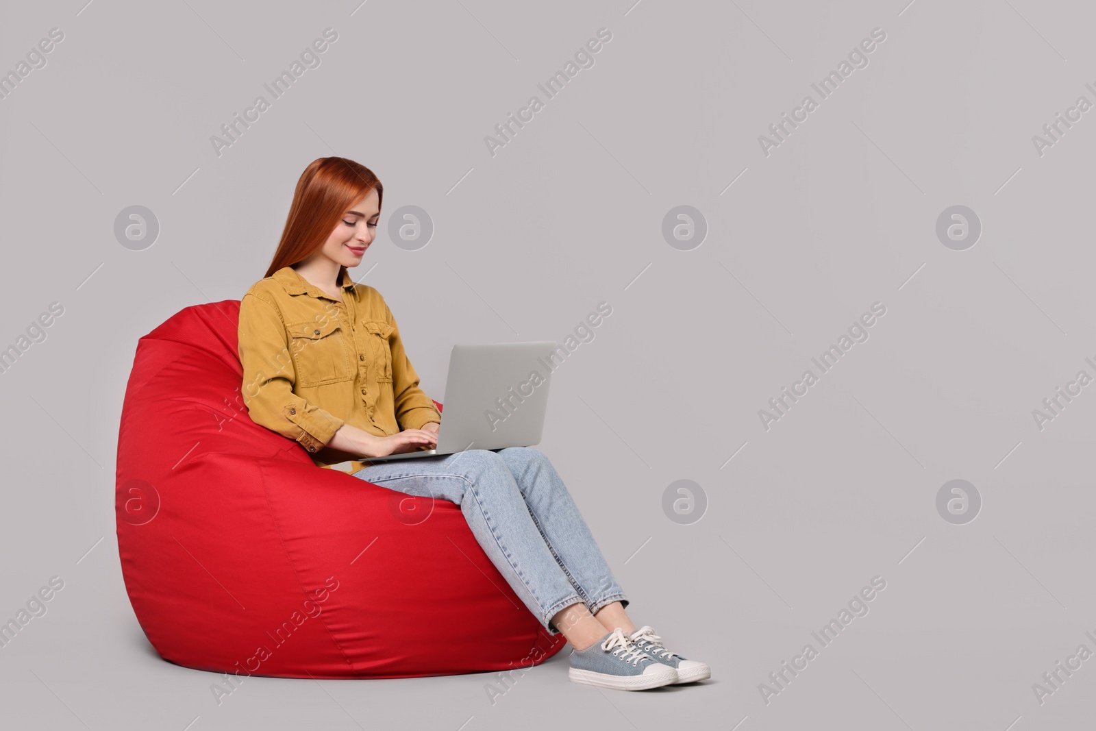 Photo of Smiling young woman working with laptop on beanbag chair against grey background, space for text