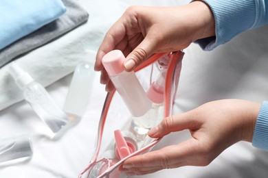 Photo of Cosmetic travel kit. Woman putting small bottle with personal care product into plastic bag on bed, closeup