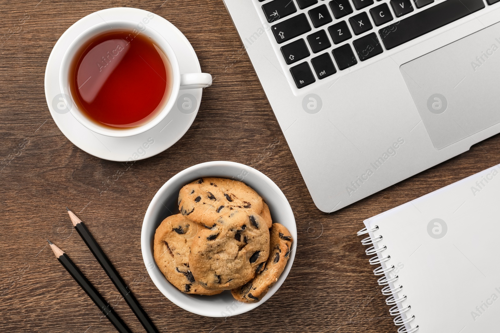 Photo of Chocolate chip cookies, cup of tea, office supplies and laptop on wooden table, flat lay