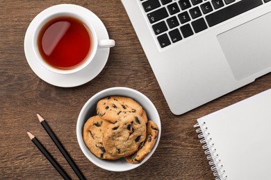 Chocolate chip cookies, cup of tea, office supplies and laptop on wooden table, flat lay