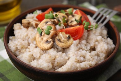 Photo of Delicious barley porridge with mushrooms, tomatoes and microgreens in bowl on table, closeup