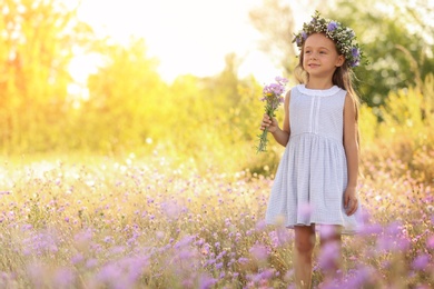 Photo of Cute little girl wearing beautiful wreath with bouquet of wildflowers outdoors, space for text. Child spending time in nature