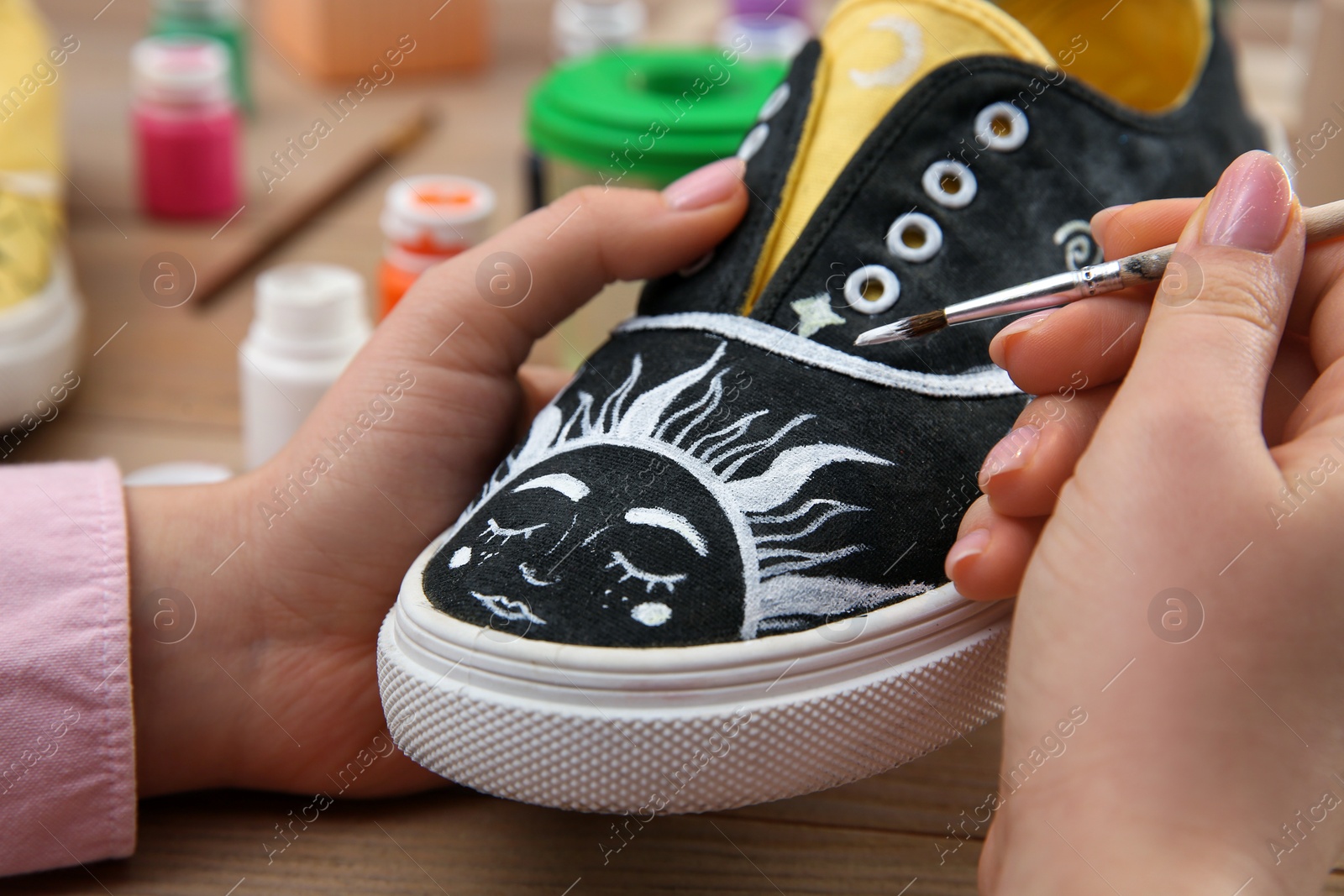 Photo of Woman painting on sneaker at wooden table, closeup. Customized shoes