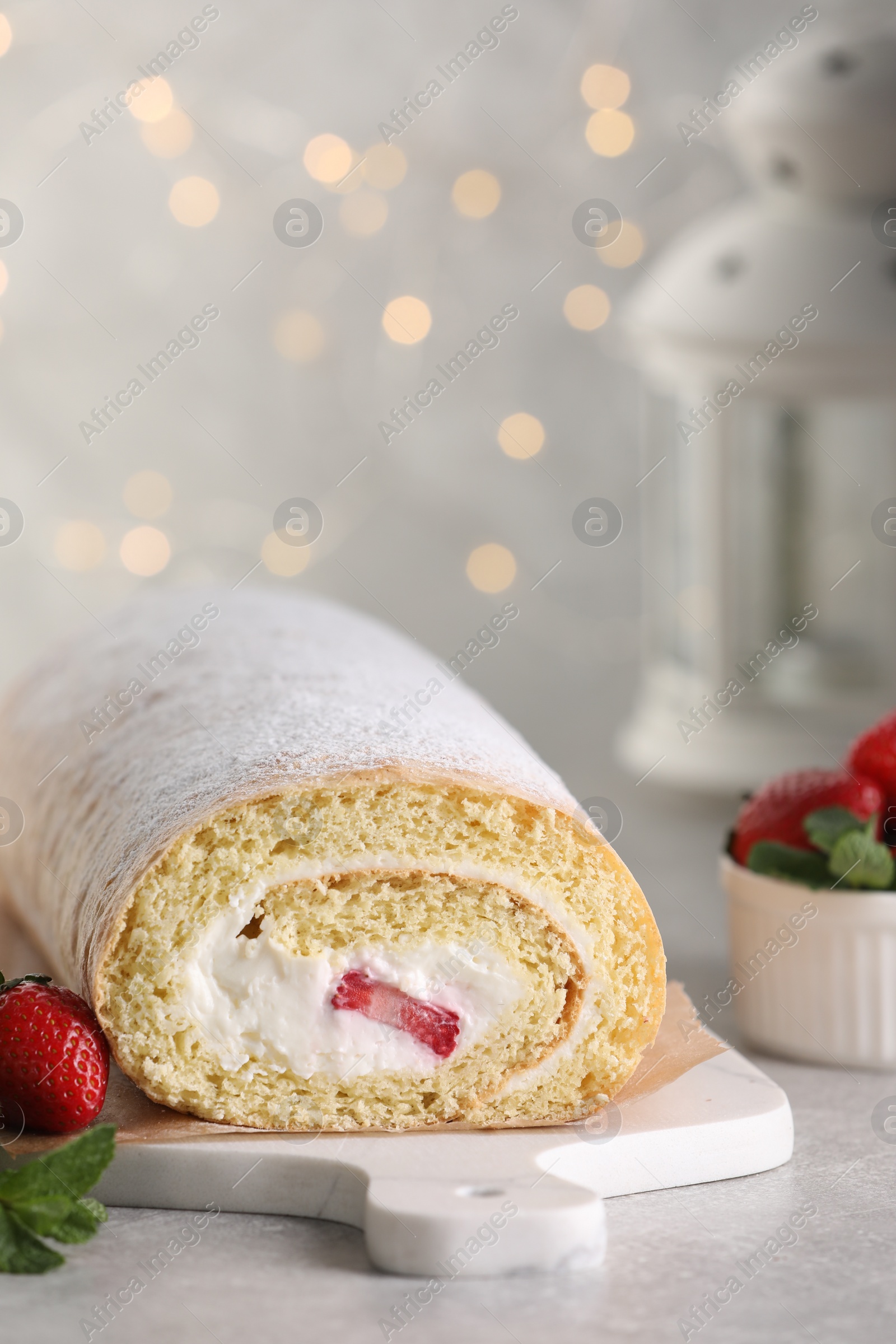 Photo of Delicious sponge cake roll with strawberries and cream on light grey table against blurred lights, space for text