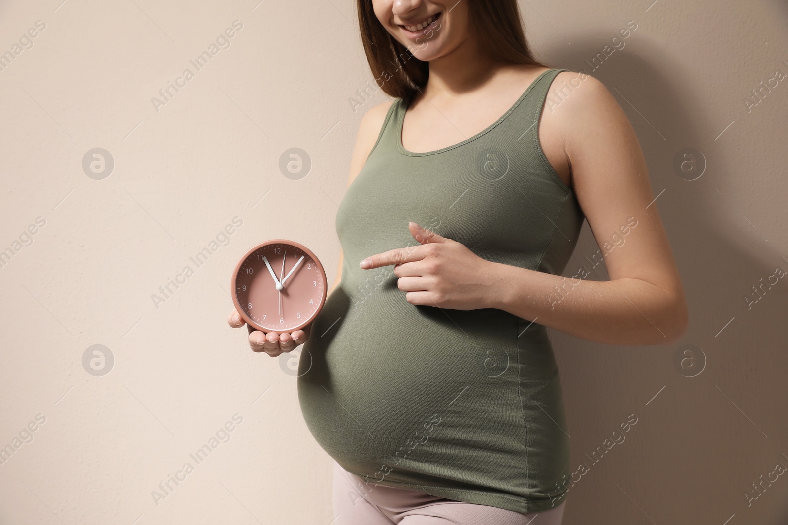 Photo of Young pregnant woman pointing at clock near her belly on beige background, closeup. Time to give birth