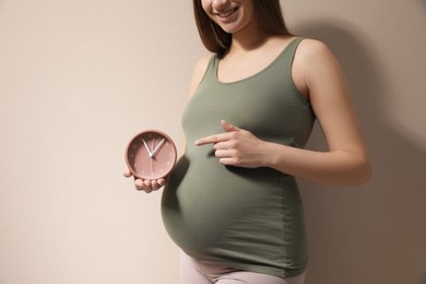 Young pregnant woman pointing at clock near her belly on beige background, closeup. Time to give birth