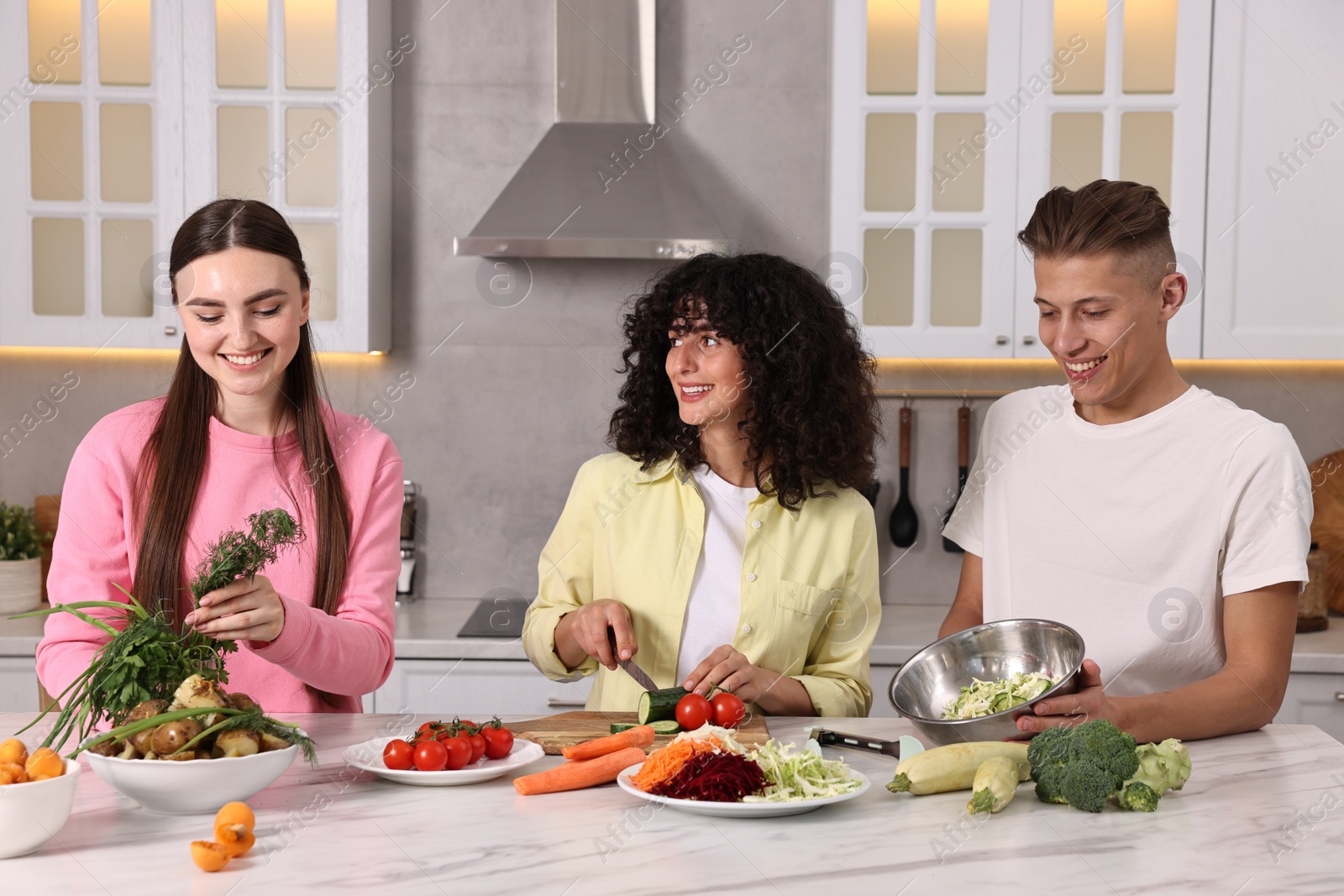 Photo of Friends cooking healthy vegetarian meal at white marble table in kitchen