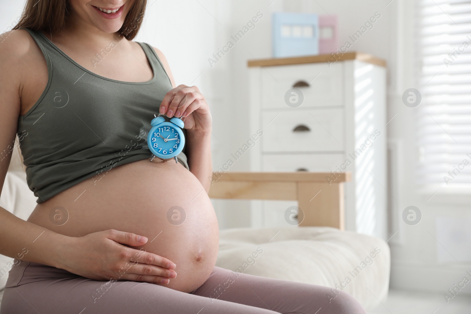 Photo of Young pregnant woman holding alarm clock near her belly at home, closeup. Time to give birth