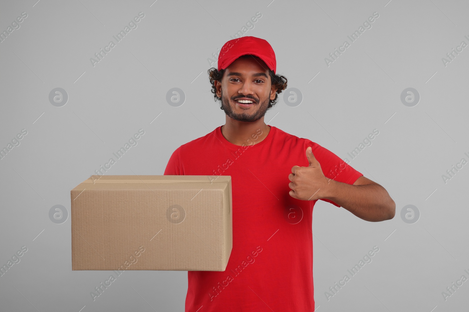 Photo of Happy young courier with parcel showing thumb up on grey background