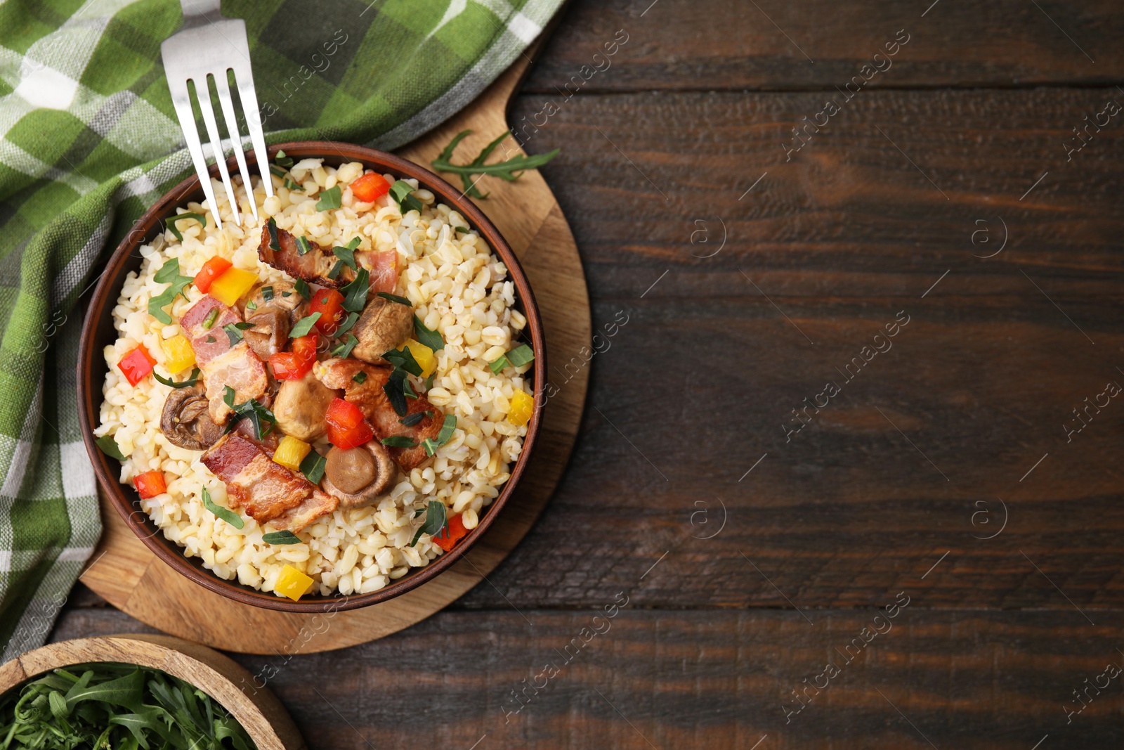 Photo of Cooked bulgur with vegetables, fried bacon and mushrooms in bowl on wooden table, top view. Space for text