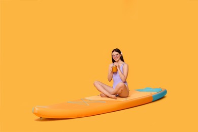 Photo of Happy woman with refreshing drink resting on SUP board against orange background