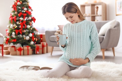 Photo of Happy pregnant woman with cup of tea sitting on floor in room decorated for Christmas