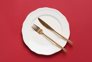 Photo of Clean plate with golden cutlery on red background, top view