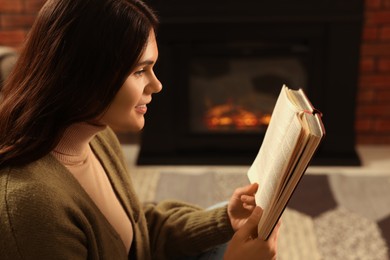 Photo of Young woman reading book on sofa near fireplace at home