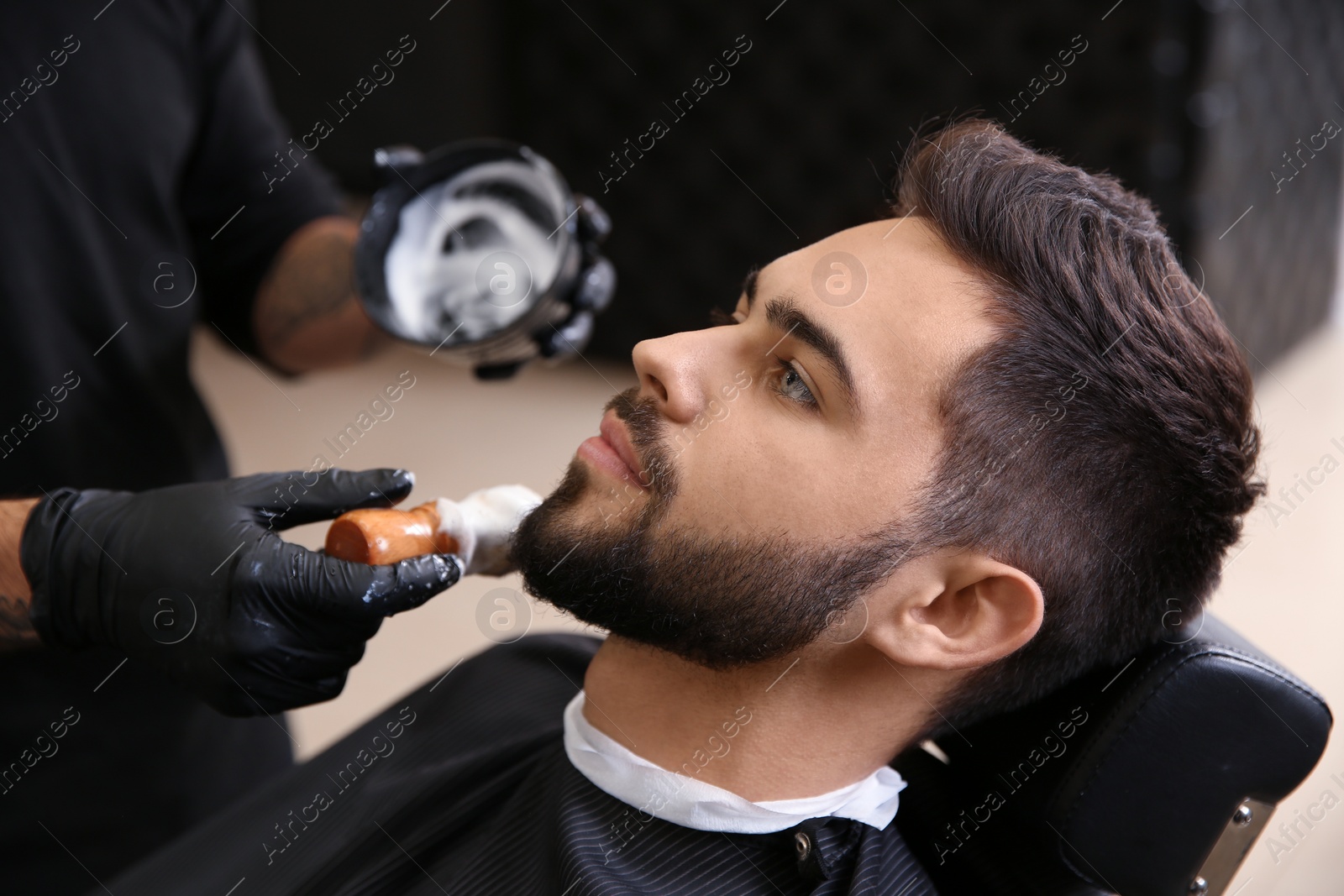 Photo of Professional hairdresser applying shaving foam onto client's skin in barbershop