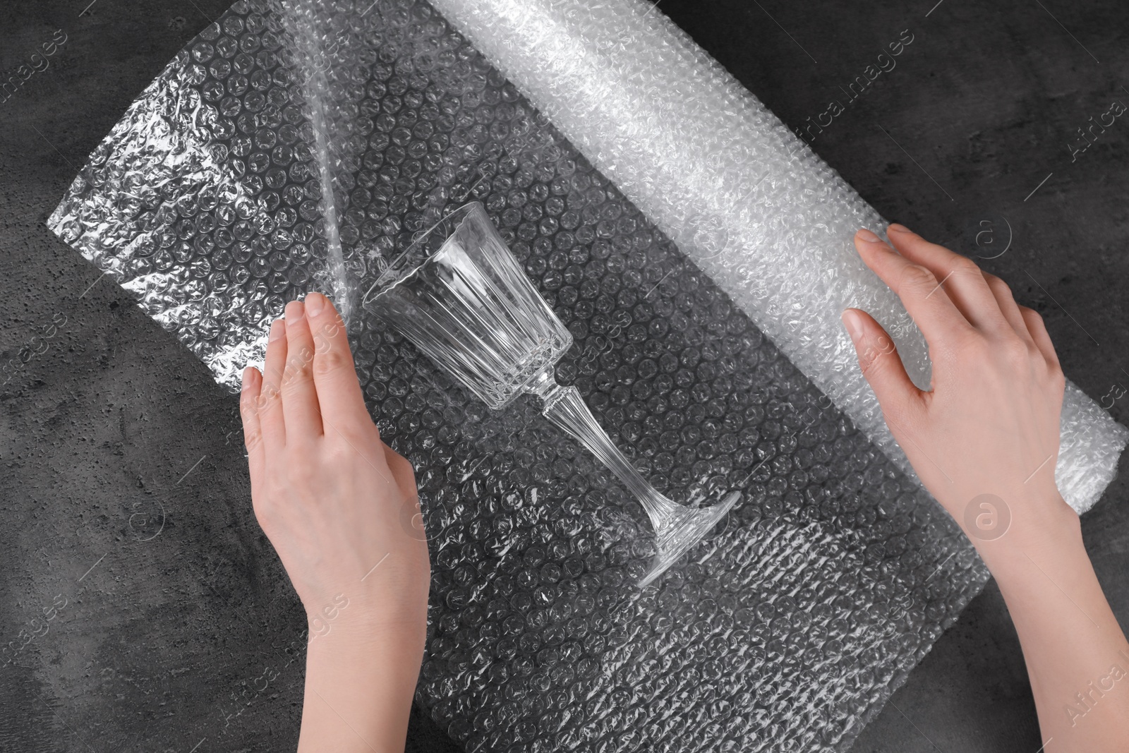 Photo of Woman covering wineglass with bubble wrap at dark grey table, closeup