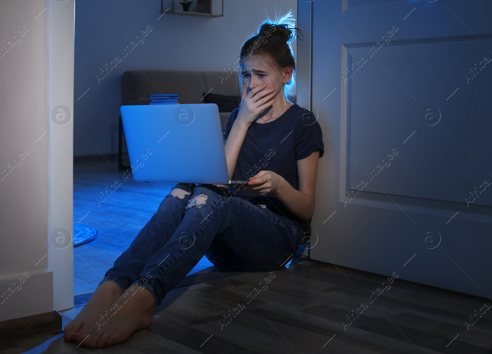 Photo of Frightened teenage girl with laptop on floor in dark room. Danger of internet