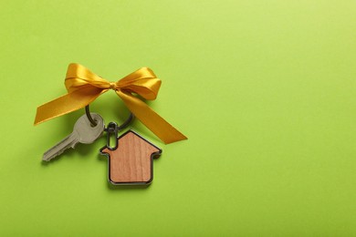 Photo of Key with trinket in shape of house and yellow bow on light green background, top view. Space for text. Housewarming party