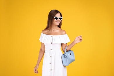 Young woman wearing stylish white dress with elegant bag on yellow background