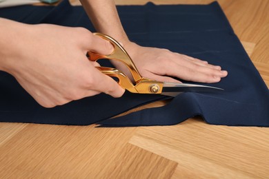 Photo of Man cutting blue fabric with scissors at wooden table, closeup