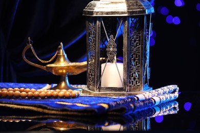 Photo of Arabic lantern, misbaha, Aladdin magic lamp and folded prayer mat on mirror surface against blurred lights at night