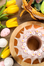 Photo of Delicious Easter cake decorated with sprinkles near painted eggs and tulips on wooden table, flat lay