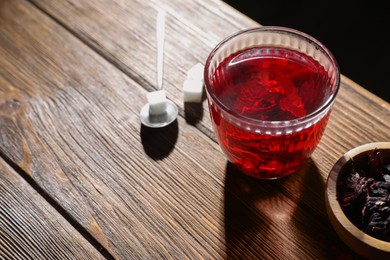 Delicious hibiscus tea in glass, sugar cubes and dry roselle petals on wooden table, above view. Space for text