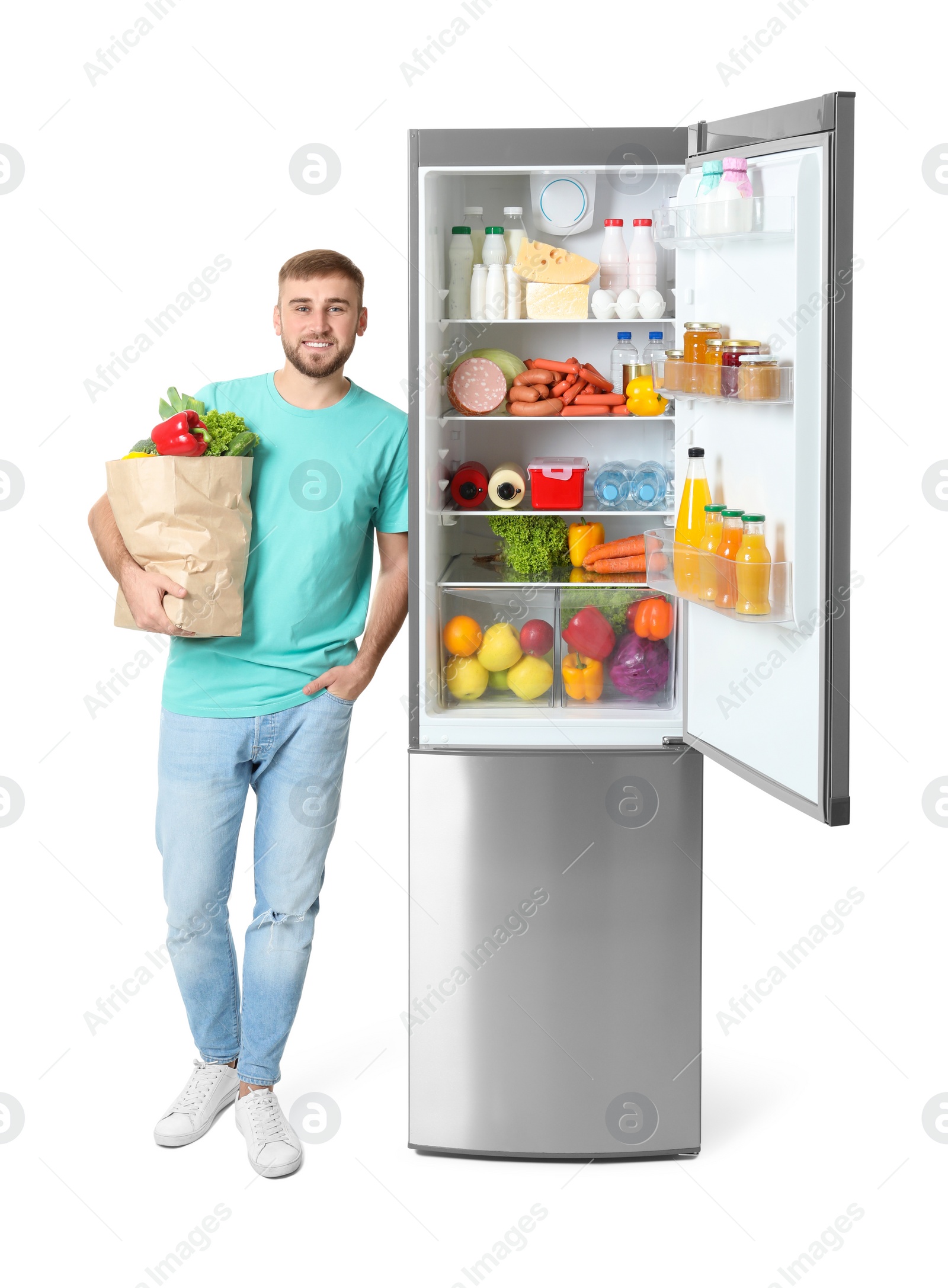 Photo of Young man with bag of groceries near open refrigerator on white background