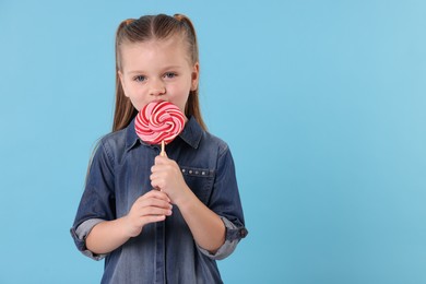 Photo of Cute little girl licking bright lollipop swirl on light blue background, space for text