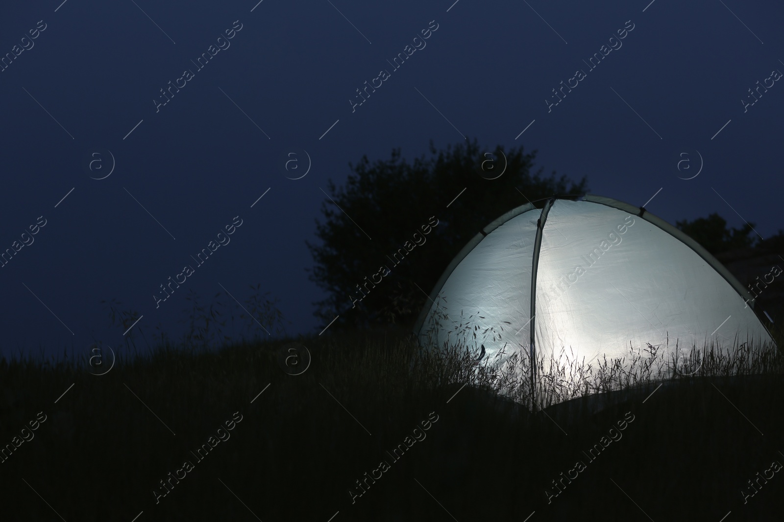 Photo of Modern tent lit from inside in wilderness at night, space for text. Overnight camping
