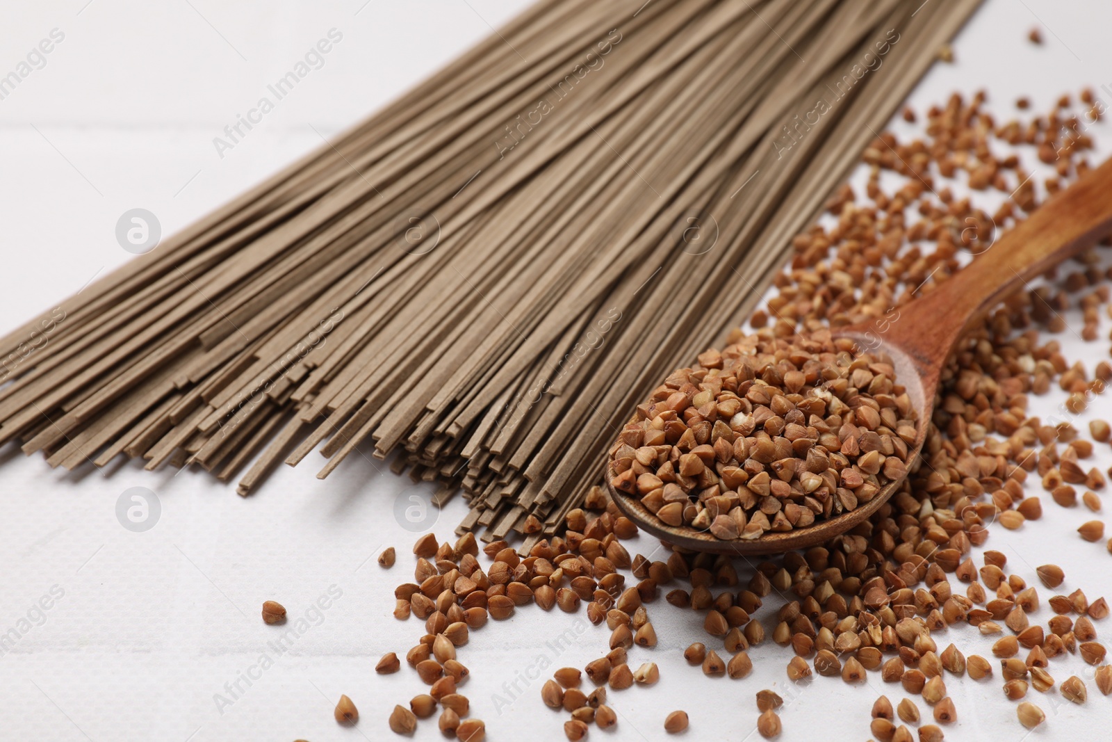 Photo of Uncooked buckwheat noodles (soba), grains and spoon on white table, closeup