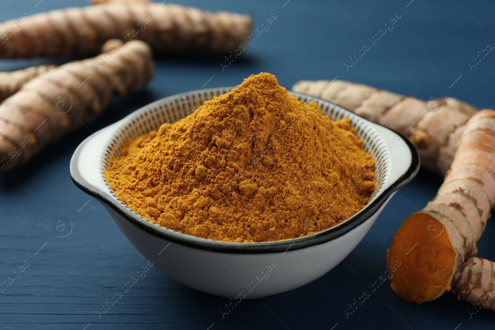 Photo of Bowl with turmeric powder and raw roots on blue wooden table, closeup