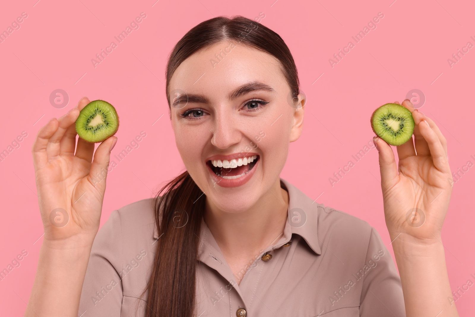 Photo of Smiling woman holding halves of kiwi on pink background