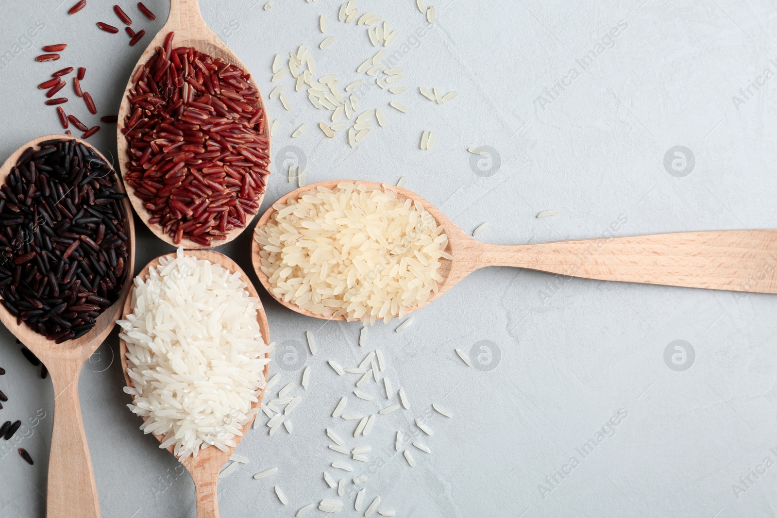 Photo of Flat lay composition with brown and other types of rice in wooden spoons on grey background