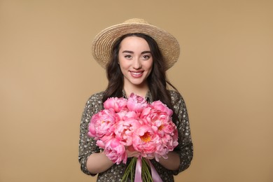 Beautiful young woman in straw hat with bouquet of pink peonies against light brown background