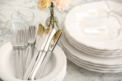 Photo of Stacked plates, cutlery and glasses on white marble table, closeup